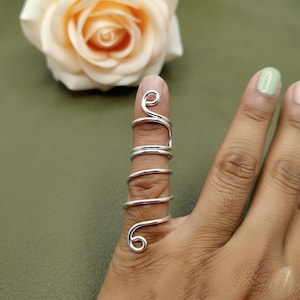 May include: A silver-colored finger ring with a unique, swirling design. The ring wraps around the finger with a series of loops and curves, creating an artistic and eye-catching accessory. The ring is shown on a finger with a light green nail polish.