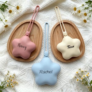 May include: Three star-shaped silicone pacifier clips with attached loops. One is dusty pink with the name Nancy, one is cream with the name Tessa, and one is light blue with the name Rachel. Each clip features a small star detail on the strap.