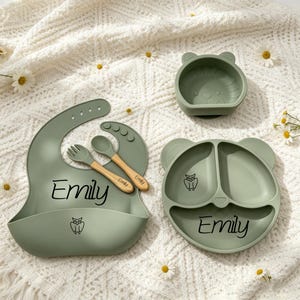 May include: A sage green silicone feeding set featuring a bib, a three-section bear-shaped plate, a small bowl, and a fork and spoon with wooden handles. The bib and plate are personalised with the name Emily and a small owl graphic.