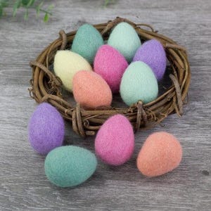 May include: A collection of pastel-colored felt Easter eggs in a woven twig nest. The eggs are in shades of pink, purple, yellow, and green. The nest sits on a weathered gray wooden surface, with greenery in the background.