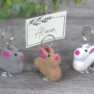 May include: Three felt bunny place card holders with pink ears and noses. The bunny on the right is white, the middle bunny is brown, and the bunny on the left is gray. Each bunny is attached to a silver spring.