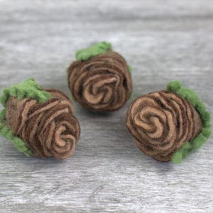 May include: Three small, round, needle-felted wool objects shaped like roses. Each piece features a spiraled center in shades of light and dark brown, surrounded by a ruffled, textured outer layer of bright green wool fibers.