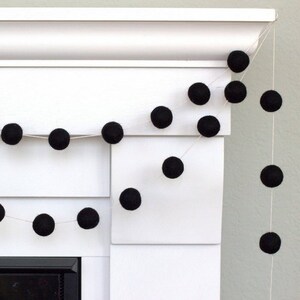 May include: A decorative garland consisting of numerous solid black felt balls strung together on a thin white cord. The spheres are evenly spaced along the string, creating a repeating pattern of dark circular shapes against the white background.