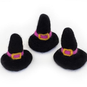 May include: Three miniature black witch hats with pink bands and gold buckles are arranged on a white background. These felt hats are a festive decoration for Halloween or other themed events. The hats are small and detailed.