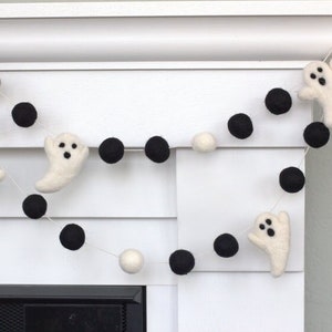 May include: A white and black felt garland with white ghost shapes and black pom poms. The garland is hanging on a white wall.