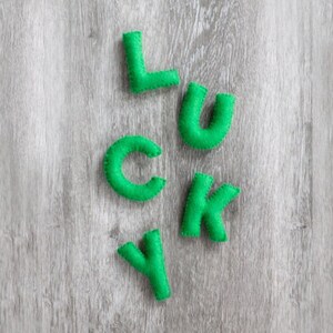 May include: Bright green felt letters spell out the word "LUCKY" against a light gray wood grain background. The letters are hand-stitched and have a soft, textured appearance. The letters are evenly spaced and arranged in a playful manner.