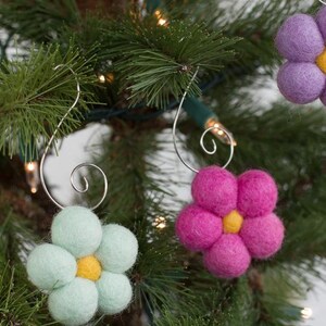May include: Close-up of a Christmas tree decorated with felt flower ornaments. The ornaments are in shades of pink, purple, and light green, each with a yellow center. The ornaments are suspended by silver wire hooks.
