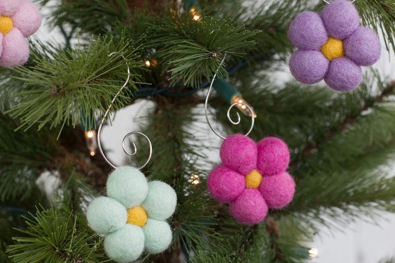 May include: Close-up of a Christmas tree decorated with felt flower ornaments. The ornaments are in shades of pink, purple, and light green, each with a yellow center. The ornaments are suspended by silver wire hooks.