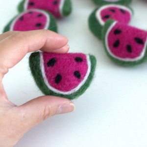 May include: A felt watermelon slice with a green rind, pink flesh, and black seeds. The slice is being held in a hand.