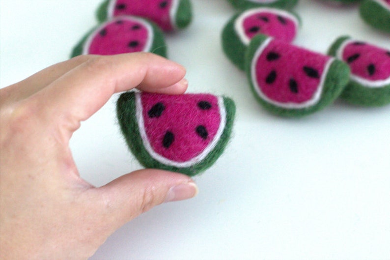 May include: A felt watermelon slice with a green rind, pink flesh, and black seeds. The slice is being held in a hand.