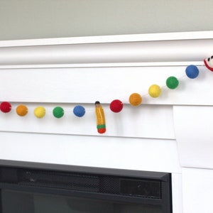 May include: A colorful felt ball garland with red apple, yellow, orange, green, blue, and red felt balls. A felt pencil is hanging from the garland. The garland is hanging above a white fireplace mantel.
