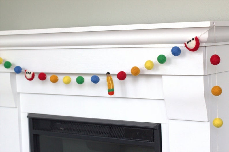 May include: A colorful felt ball garland with red apple, yellow, orange, green, blue, and red felt balls. A felt pencil is hanging from the garland. The garland is hanging above a white fireplace mantel.