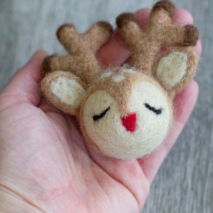 May include: A handcrafted felt reindeer head with closed eyes and a red nose. The reindeer has brown antlers and ears, and is held in a person's hand. The reindeer is a charming, whimsical decoration.