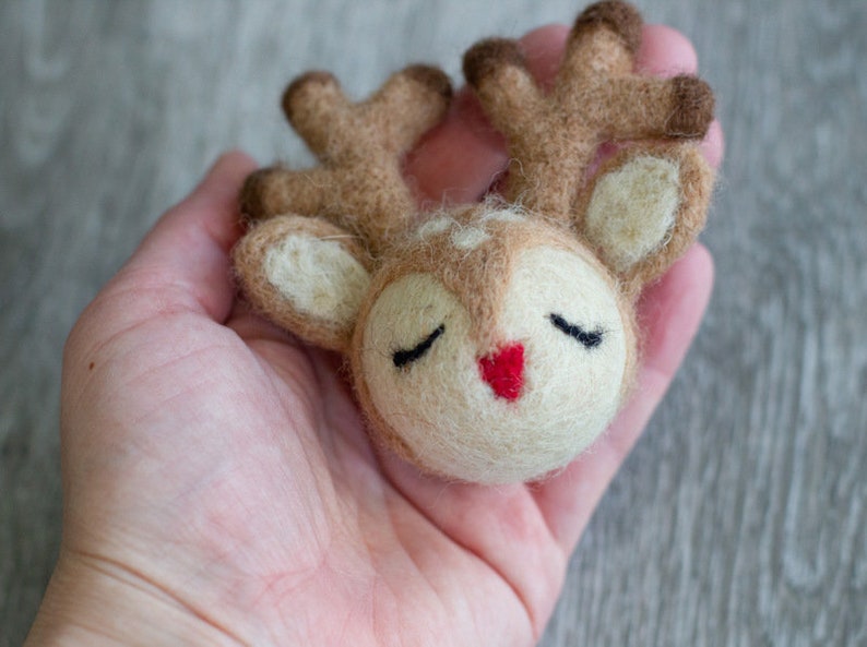 May include: A handcrafted felt reindeer head with closed eyes and a red nose. The reindeer has brown antlers and ears, and is held in a person's hand. The reindeer is a charming, whimsical decoration.