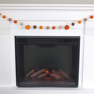 May include: A decorative garland featuring alternating felt balls in shades of orange, brown, and beige, interspersed with small orange felt pumpkins. The garland is strung on a thin white cord and hangs in a draped pattern across a white fireplace.