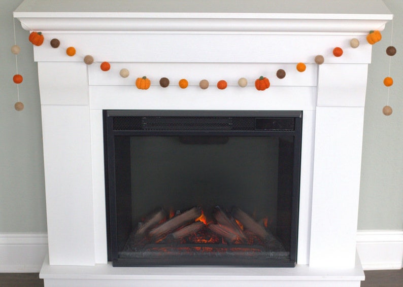 May include: A decorative garland featuring alternating felt balls in shades of orange, brown, and beige, interspersed with small orange felt pumpkins. The garland is strung on a thin white cord and hangs in a draped pattern across a white fireplace.
