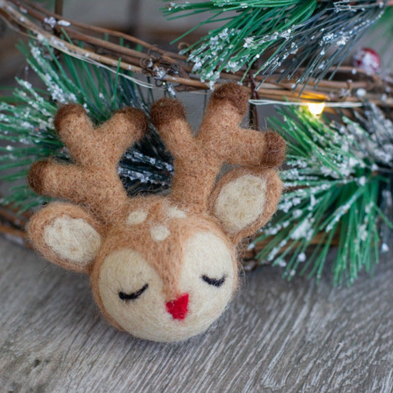 May include: A handcrafted felt reindeer ornament with closed eyes and a red nose. The reindeer has brown antlers and is set against a backdrop of a Christmas wreath with green pine needles and white accents. A charming holiday decoration.