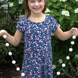 May include: A girl wearing a floral dress and a headband holds a white pom-pom garland. The garland is 4 feet long and has 16 balls. Featured Garland: 4 ft &hearts; 16 balls