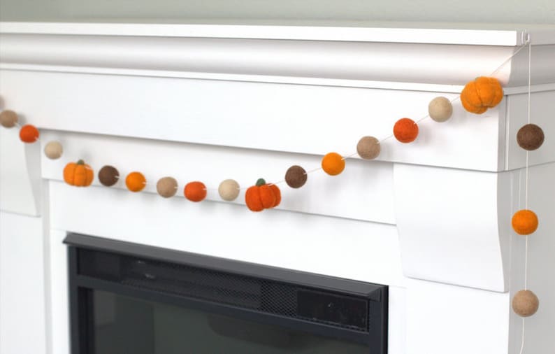 May include: A decorative garland featuring alternating felted wool balls and small pumpkin shapes in shades of orange, brown, and beige. The felted elements are strung together on a thin white cord, creating a repeating pattern of spheres and pumpkins.