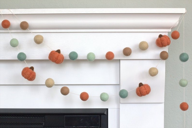 May include: A garland of felt balls in shades of green, brown, and orange with felt pumpkin accents. The garland is strung on a white background.
