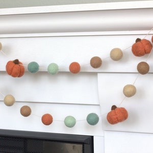 May include: A garland of felt balls in shades of brown, green, and orange, with felt pumpkin shapes interspersed throughout. The garland is strung on a white string and is hanging from a white mantel.