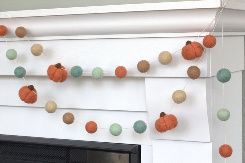 May include: A garland of felt balls in shades of brown, green, and orange, with felt pumpkin shapes interspersed throughout. The garland is strung on a white string and is hanging from a white mantel.