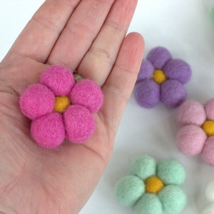 May include: A close-up of several felt flowers in various colors, including pink, purple, green, and white. The flowers have a yellow center and are made of soft, textured material. One pink flower is held in a hand.