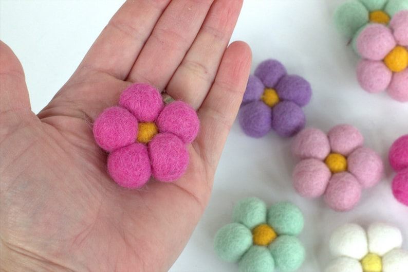 May include: A close-up of several felt flowers in various colors, including pink, purple, green, and white. The flowers have a yellow center and are made of soft, textured material. One pink flower is held in a hand.