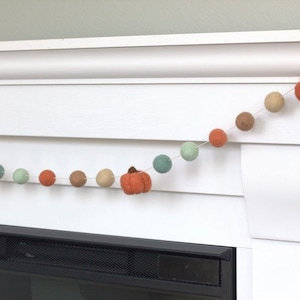 May include: A garland of felt balls in shades of green, brown, and orange with small felt pumpkins hanging between the balls. The garland is strung across a white fireplace mantel.