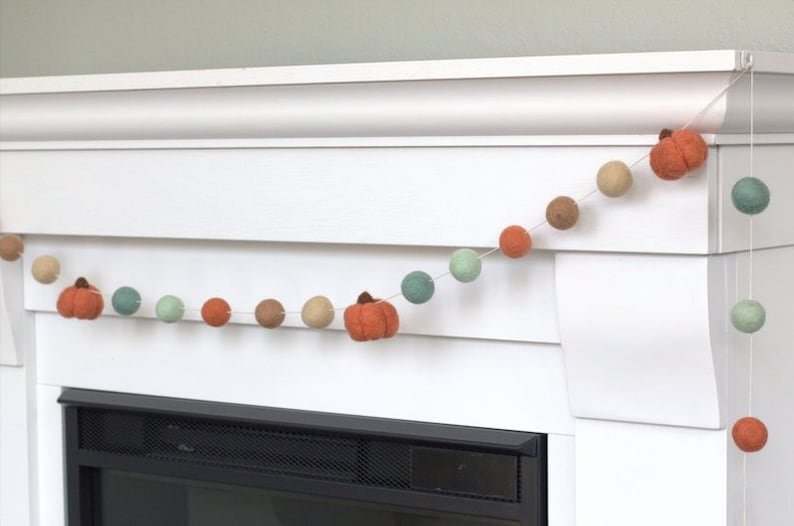 May include: A garland of felt balls in shades of green, brown, and orange with small felt pumpkins hanging between the balls. The garland is strung across a white fireplace mantel.