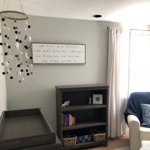 May include: A nursery room with a white crib, a wooden bookshelf, a white armchair with a blue throw blanket, and a white curtain with black beads. A wooden sign with the text "I'll love you forever. I'll like you for always. As long as I'm living, my baby you'll be." hangs on the wall. A white and gray pom-pom mobile hangs from the ceiling.