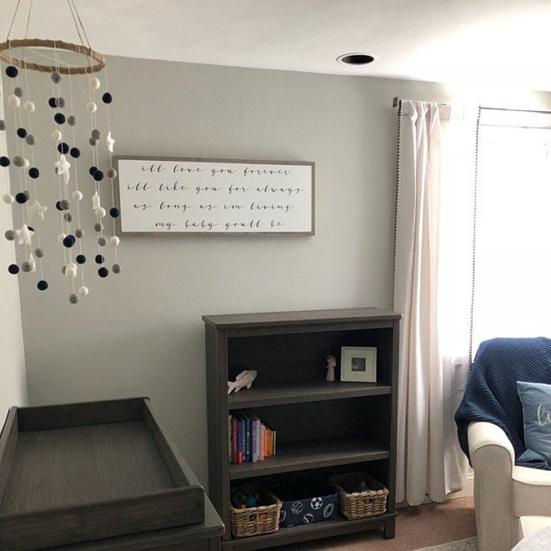 May include: A nursery room with a white crib, a wooden bookshelf, a white armchair with a blue throw blanket, and a white curtain with black beads. A wooden sign with the text "I'll love you forever. I'll like you for always. As long as I'm living, my baby you'll be." hangs on the wall. A white and gray pom-pom mobile hangs from the ceiling.