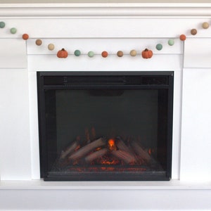 May include: A white fireplace mantel with a garland of felt pumpkins and balls in shades of orange, brown, and green. The fireplace has a black electric insert with a realistic flame effect.