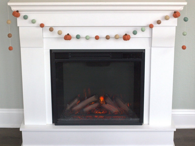 May include: A white fireplace mantel with a garland of felt pumpkins and balls in shades of orange, brown, and green. The fireplace has a black electric insert with a realistic flame effect.