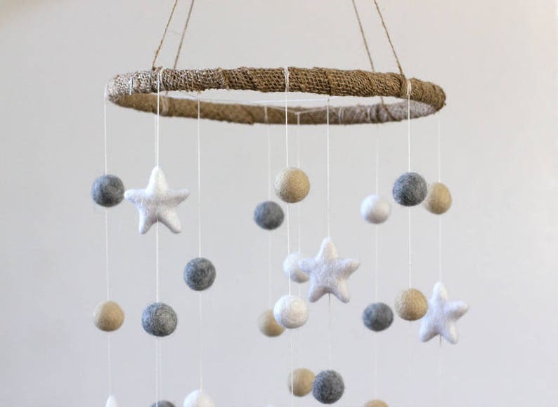 May include: A white, gray, and beige felt baby mobile with a natural jute ring. The mobile features felt stars and balls hanging from the ring.
