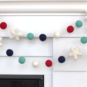 May include: A felt garland with red, white, and blue pom-poms and white felt stars. The garland is strung on a white string and is hanging on a white wall.