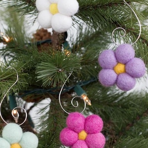 May include: Close-up of a Christmas tree decorated with handmade felt flower ornaments. The ornaments are white, purple, pink, and light green with yellow centers. Each flower has a silver wire hanger.