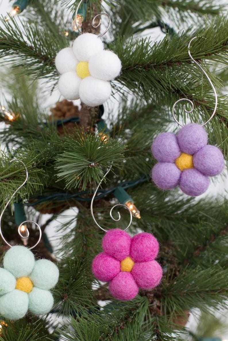 May include: Close-up of a Christmas tree decorated with handmade felt flower ornaments. The ornaments are white, purple, pink, and light green with yellow centers. Each flower has a silver wire hanger.