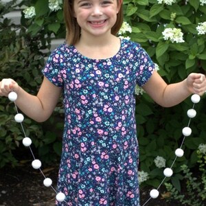 May include: A girl holds a white pom-pom garland with 16 balls. The garland is 4 feet long. The girl is wearing a blue and pink floral dress and a white headband with a flower.