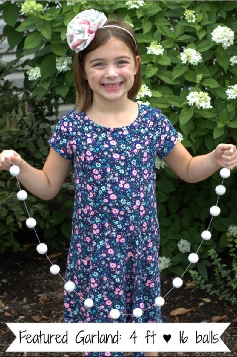 May include: A girl holds a white pom-pom garland with 16 balls. The garland is 4 feet long. The girl is wearing a blue and pink floral dress and a white headband with a flower.