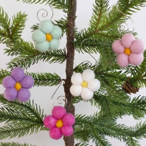 May include: A collection of colorful felt flower ornaments with yellow centers and silver wire hangers. The flowers are in shades of pink, purple, white, and light green, displayed on a small evergreen tree. A festive, handmade decoration.