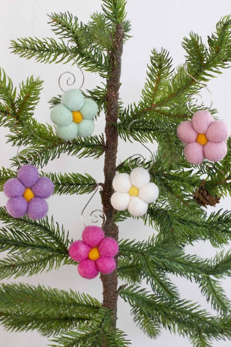May include: A collection of colorful felt flower ornaments with yellow centers and silver wire hangers. The flowers are in shades of pink, purple, white, and light green, displayed on a small evergreen tree. A festive, handmade decoration.