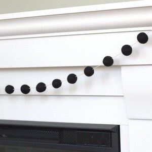 May include: A garland consisting of a thin white string adorned with evenly spaced, solid black spherical felt balls. The string is draped horizontally, showcasing a series of these dark, round, textured elements arranged in a linear sequence.