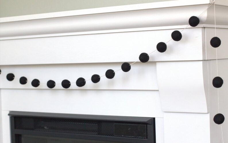 May include: A garland consisting of a thin white string adorned with evenly spaced, solid black spherical felt balls. The string is draped horizontally, showcasing a series of these dark, round, textured elements arranged in a linear sequence.