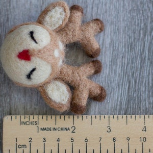 May include: A handmade felt deer with a cream-colored face, brown body, and white spots. The deer has closed eyes, a red nose, and brown antlers. A wooden ruler with inch and centimeter measurements is in the foreground.