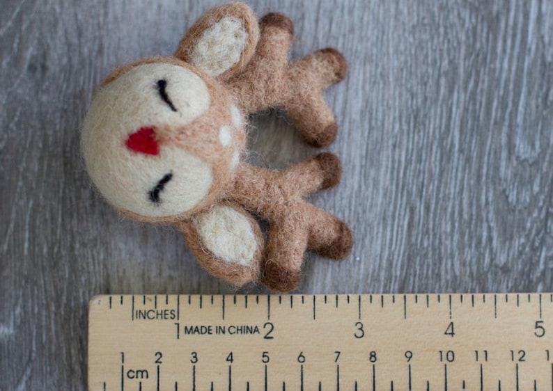 May include: A handmade felt deer with a cream-colored face, brown body, and white spots. The deer has closed eyes, a red nose, and brown antlers. A wooden ruler with inch and centimeter measurements is in the foreground.