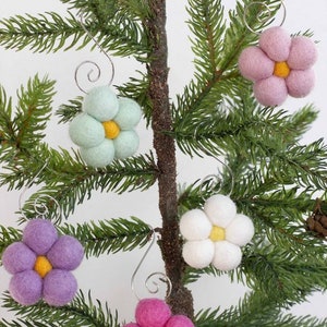 May include: Close-up of a small Christmas tree branch adorned with six felt flower ornaments. The flowers are in shades of pink, purple, white, and mint green, each with a yellow center. Silver wire hooks are attached for hanging.