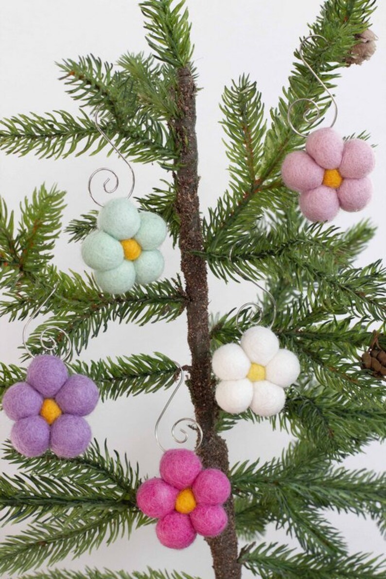 May include: Close-up of a small Christmas tree branch adorned with six felt flower ornaments. The flowers are in shades of pink, purple, white, and mint green, each with a yellow center. Silver wire hooks are attached for hanging.