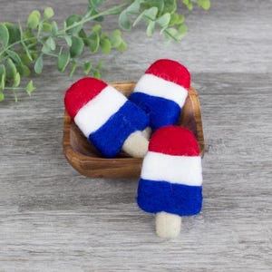 May include: Three red, white, and blue felt popsicle ornaments rest in a small wooden bowl. The popsicles have a red top, a white center, and a blue bottom, with a beige stick. The bowl sits on a gray wooden surface.
