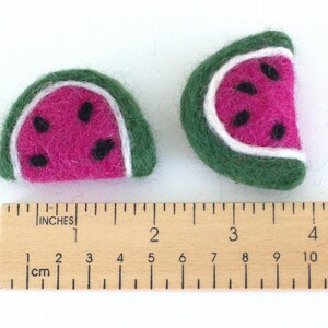 May include: Two felt watermelon slices, one larger than the other, with green rinds, white seeds, and pink flesh. The slices are measured against a wooden ruler.